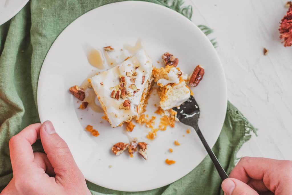 Keto pumpkin cheesecake bars on a white plate with hands with a fork taking a bite.