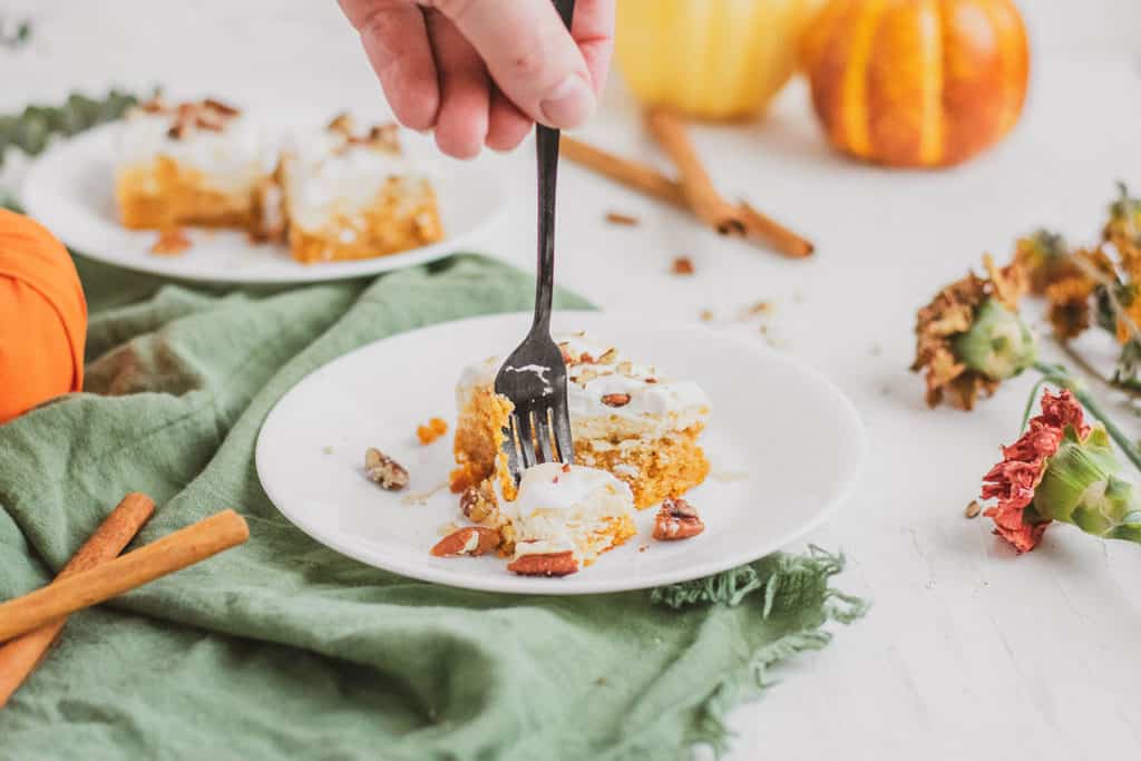 Keto pumpkin cheesecake bars on a white plate with hands with a fork taking a bite.