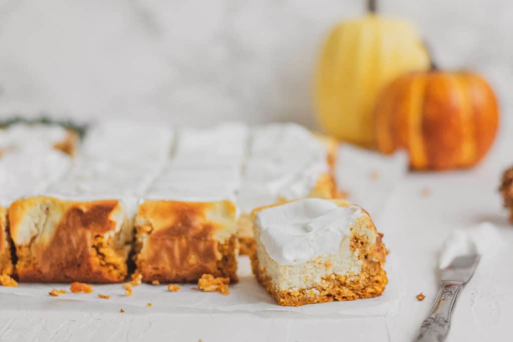 Keto pumpkin cheesecake bars on a white surface with pumpkins in the background.