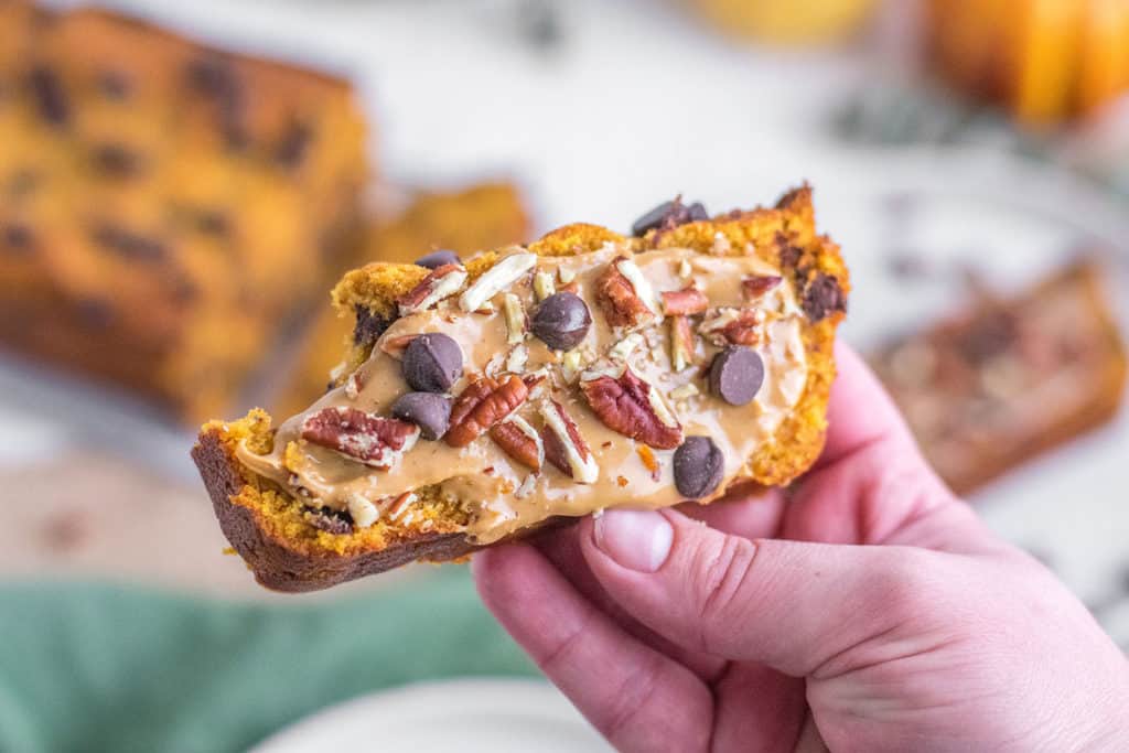 Keto Chocolate Chip Pumpkin Bread slice with nut butter and chocolate chips and pecans on top in a hand with a bite taken.