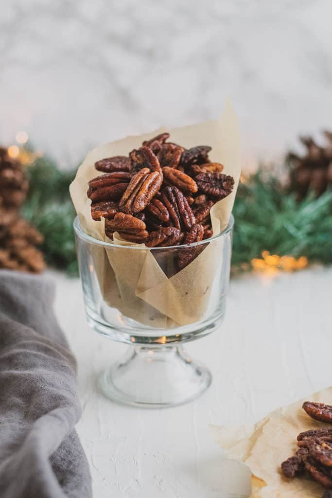 Keto Candied Pecans in a glass cup with garland and pine cones in the background on a white surface. 