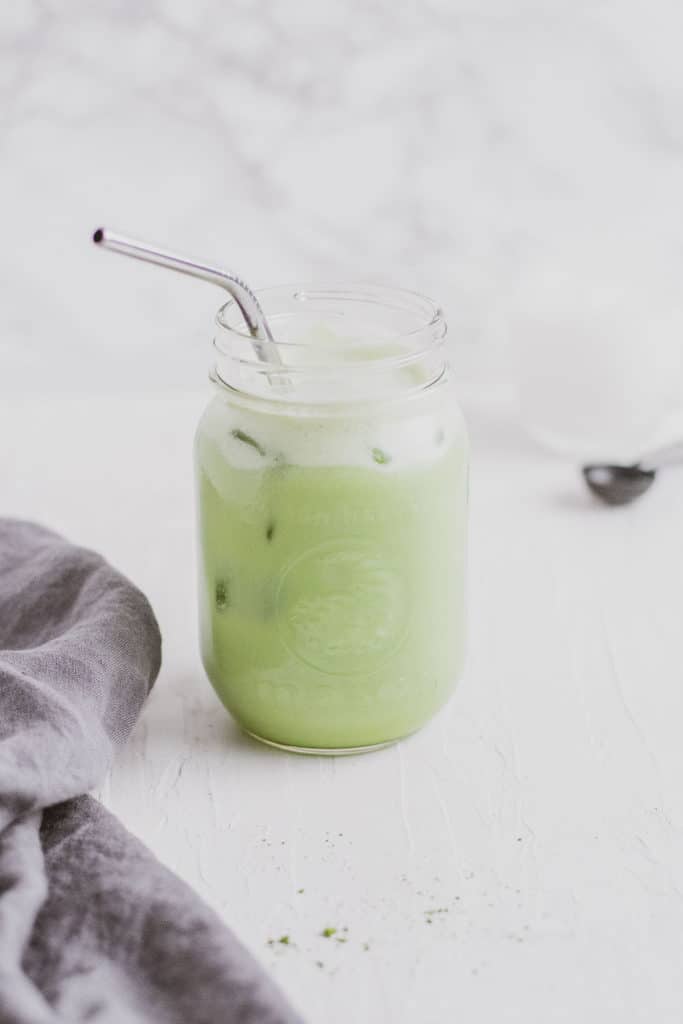 Keto iced matcha in a glass jar with a metal straw and a napkin on the side on a white surface.