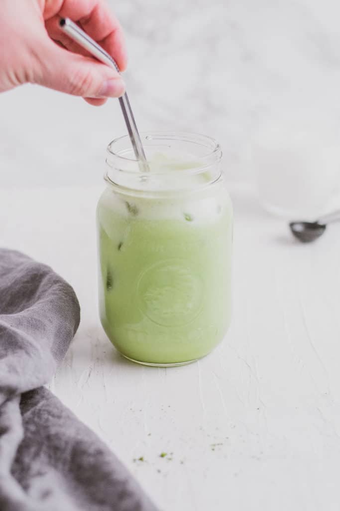 Keto iced matcha in a glass jar with a metal straw being put in by a hand and a napkin on the side on a white surface.