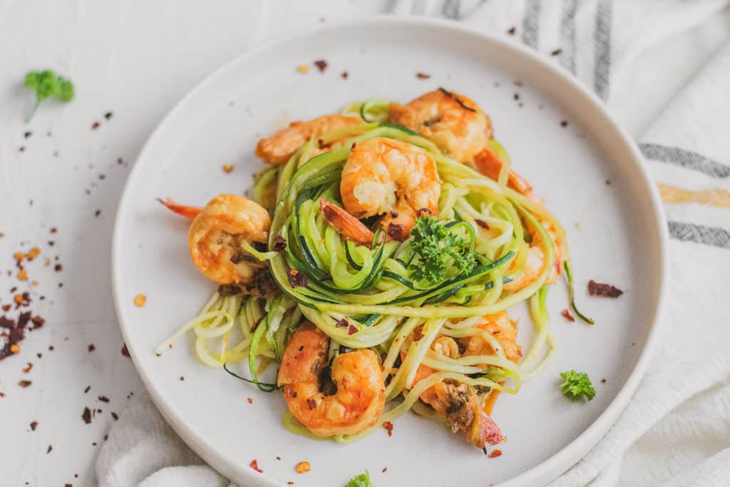 Keto shrimp scampi with zucchini noodles, shrimp, and parsley on a white plate with a  napkin on the side.