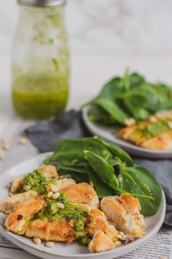Keto Chicken Pesto and basil pesto on top with a side of spinach on a white plate on newspaper with a blue napkin on the side with the bottle of pesto to the side