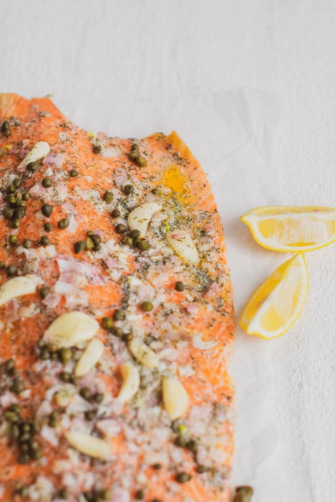 Keto Garlic Butter Baked Salmon fillet topped with butter, garlic cloves and capers on a white surface with lemon wedges on the side.
