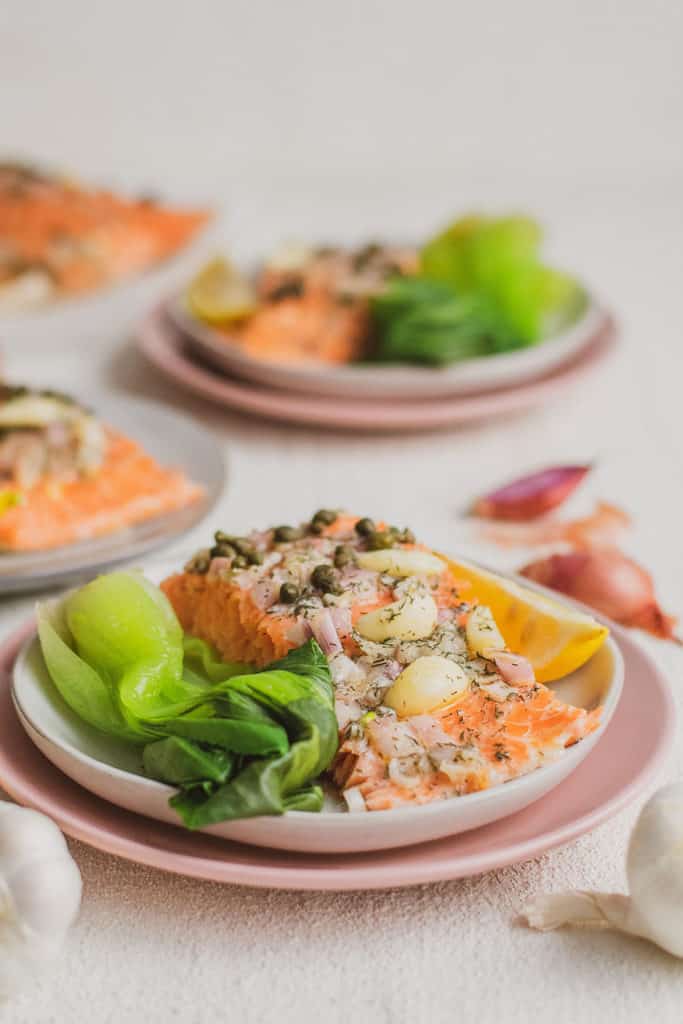Keto Garlic Butter Baked Salmon fillets on white plates and pink plates under on a white surface with a side of bok choy and a lemon wedge and shallots and garlic bulbs on the side.