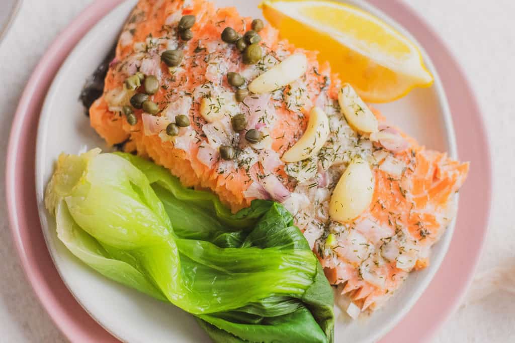 Keto Garlic Butter Baked Salmon fillets on white plates and pink plates under on a white surface with a side of bok choy and a lemon wedge and shallots and garlic bulbs on the side.