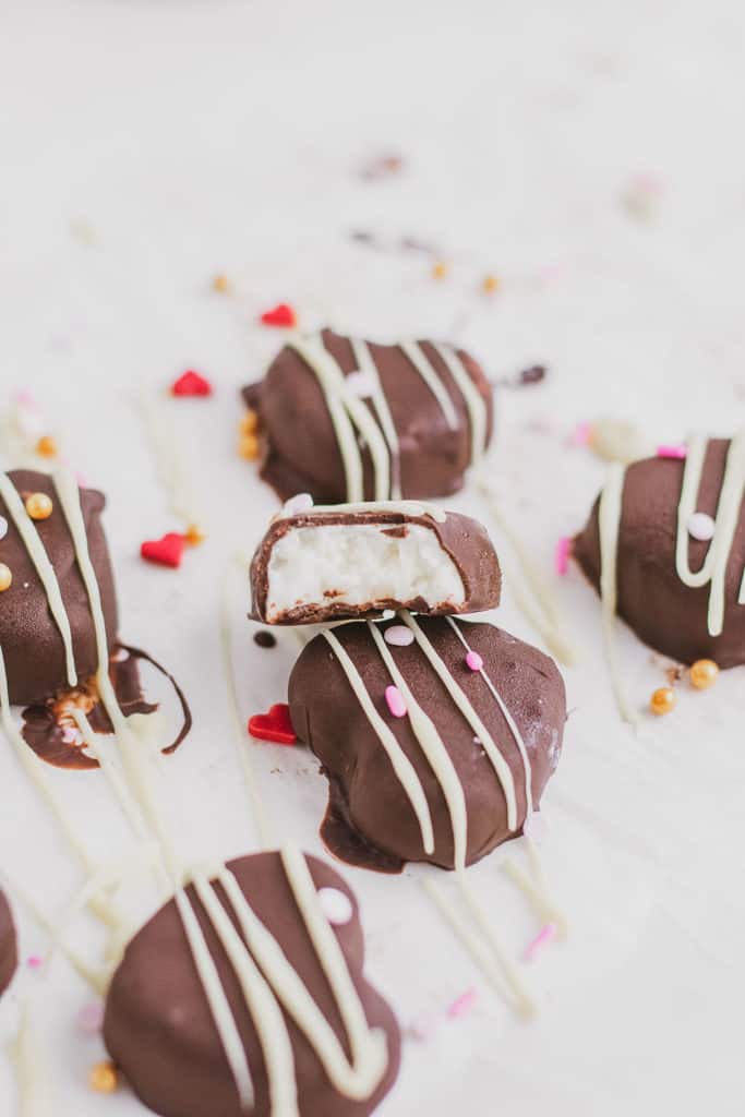 Keto Peppermint Patties shaped as a heart that are chocolate coated with white chocolate drizzle and heart sprinkles with a bite taken out on a white surface.