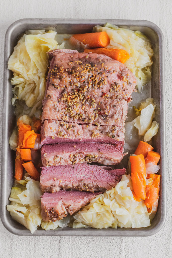 Sliced Keto Corned Beef and Cabbage on a tray with cooked carrots and onions on the side on a white surface