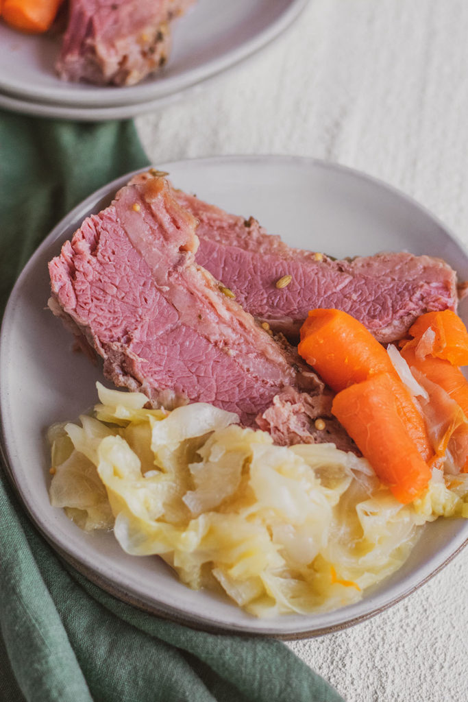 Slices Keto Corned Beef and Cabbage on a white plate with cooked carrots and onions on the side on a white surface with a green napkin on the side