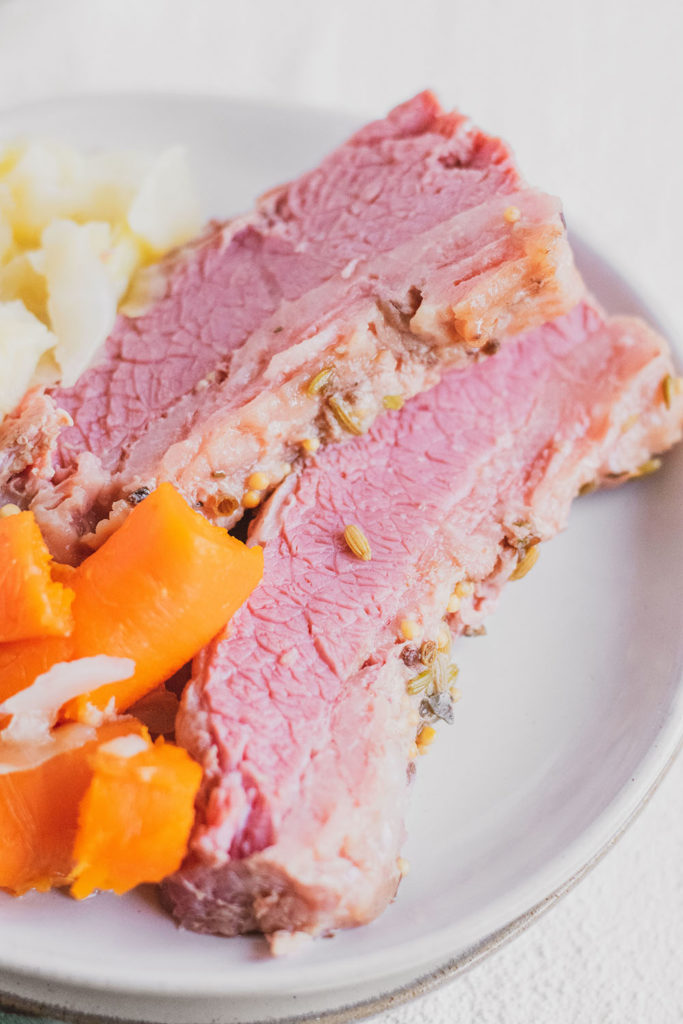 Slices Keto Corned Beef and Cabbage on a white plate with cooked carrots and onions on the side on a white surface