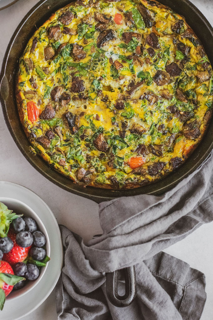 Keto Kale and Mushroom Frittata in a skillet with a blue napkin and a bowl of berries on the side