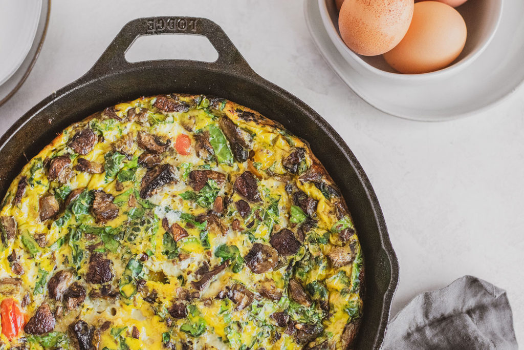 Keto Kale and Mushroom Frittata in a skillet with a blue napkin and a bowl of berries on the side
